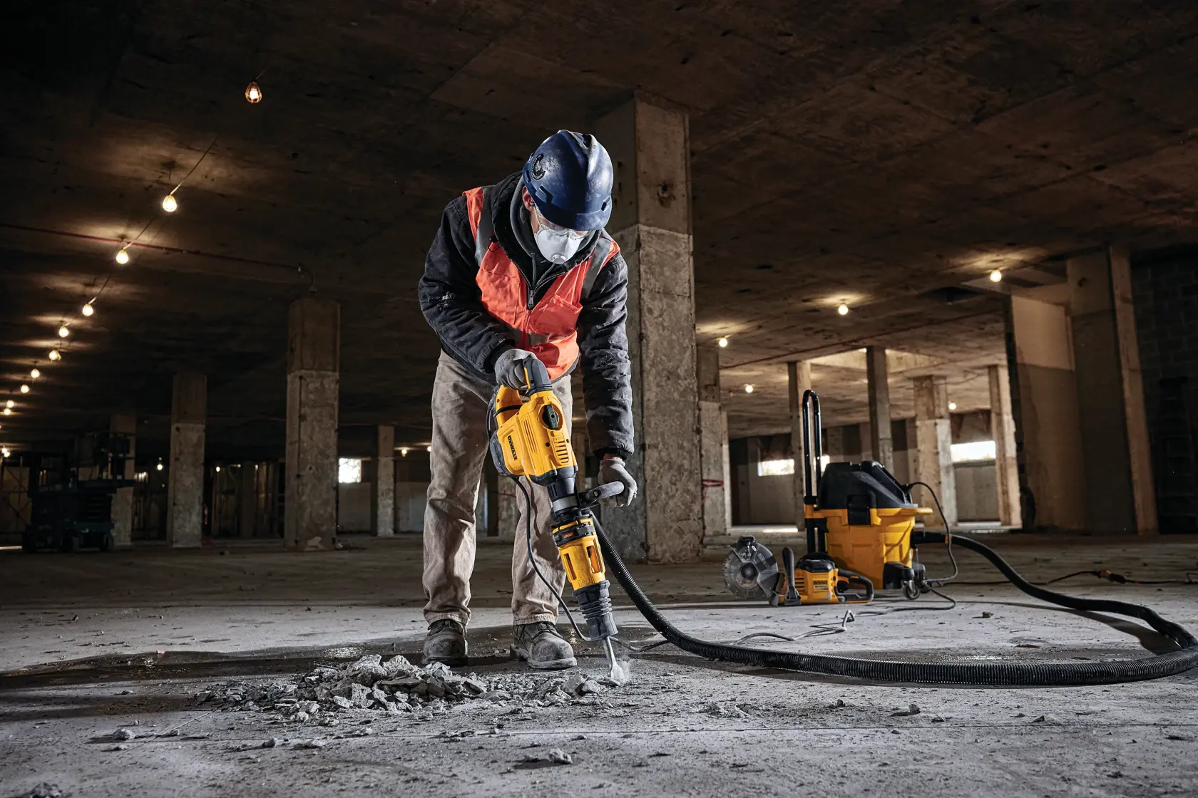 Una persona con equipo de seguridad, incluyendo casco y mascarilla, utiliza un martillo demoledor amarillo DEWALT para romper el concreto en el piso de un gran sitio de construcción. En el fondo se puede ver una máquina de aspiración amarilla DEWALT.