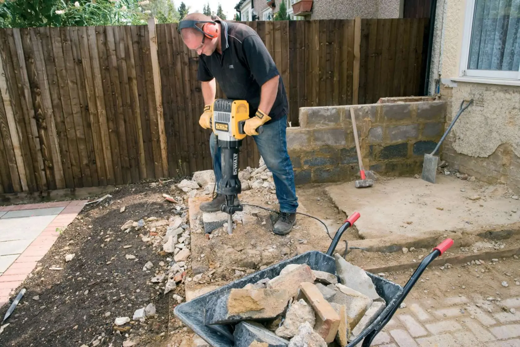 Una persona utiliza un martillo demoledor eléctrico DEWALT (modelo D25980) para romper concreto al aire libre, con una carretilla llena de escombros y piedras en primer plano.