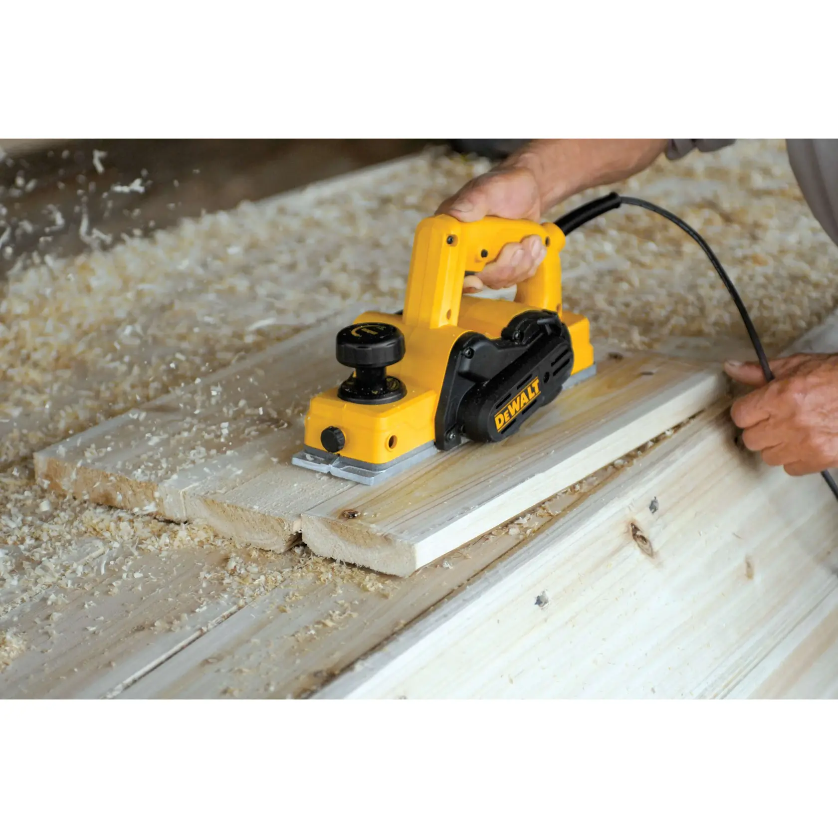Una persona usa una cepilladora eléctrica amarilla DEWALT para alisar tablas de madera, con virutas de madera dispersas alrededor del área de trabajo.
