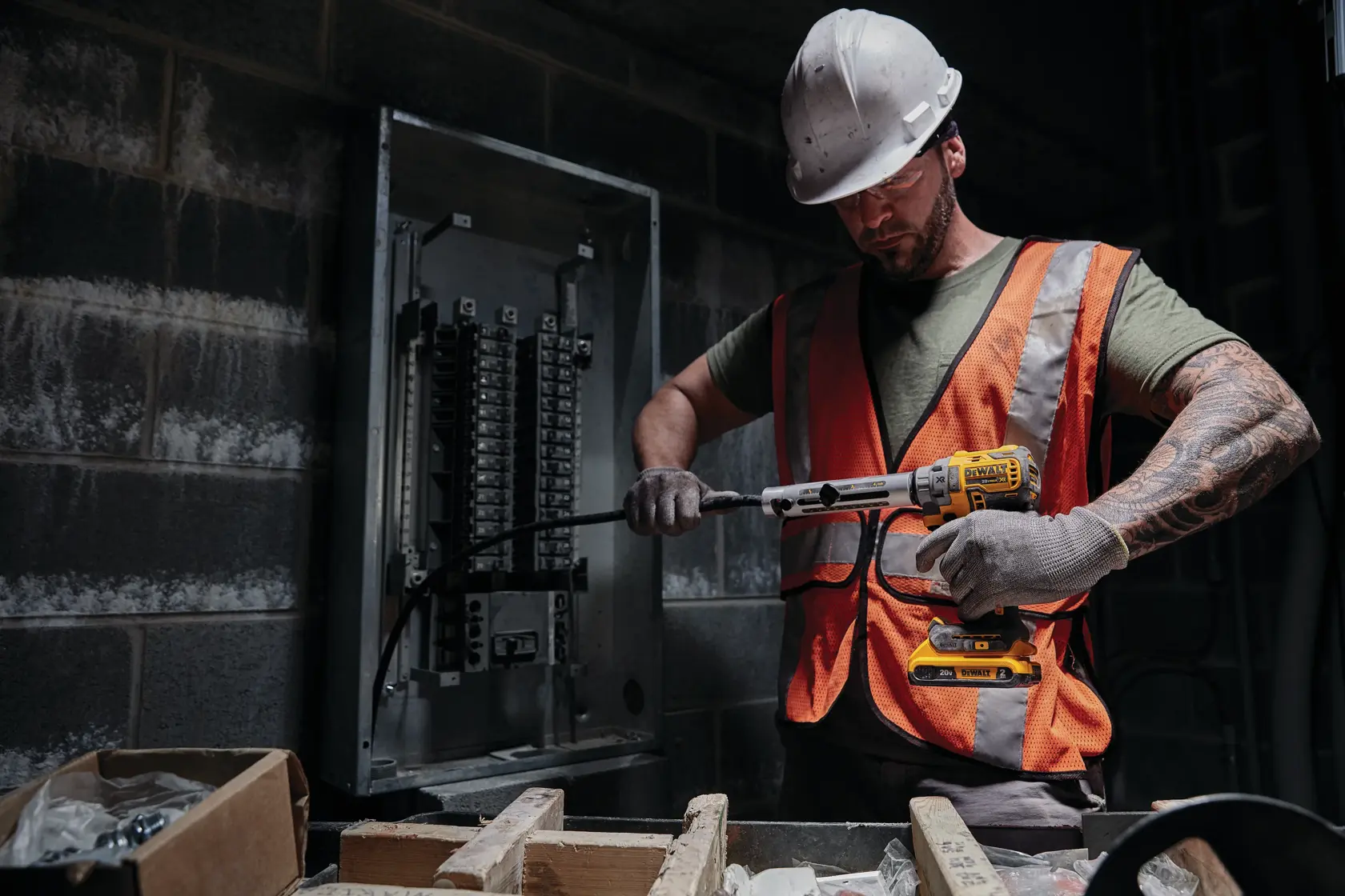 Trabajador usa taladro inalámbrico DEWALT cerca de panel eléctrico, con casco y chaleco en área industrial.