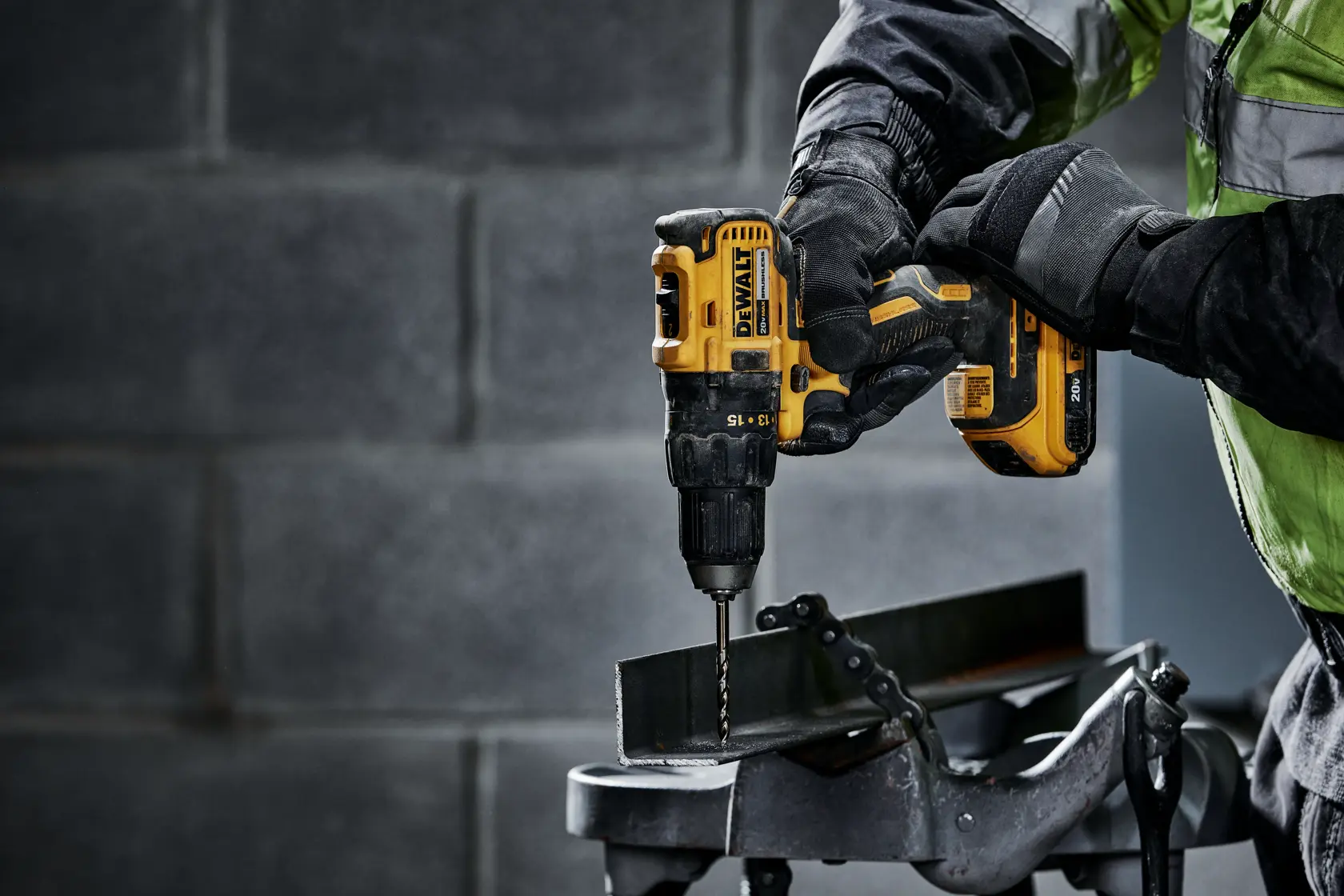 Una persona con guantes de trabajo usa un taladro inalámbrico amarillo DEWALT para perforar una pieza de metal, la cual está asegurada en una mesa de trabajo.