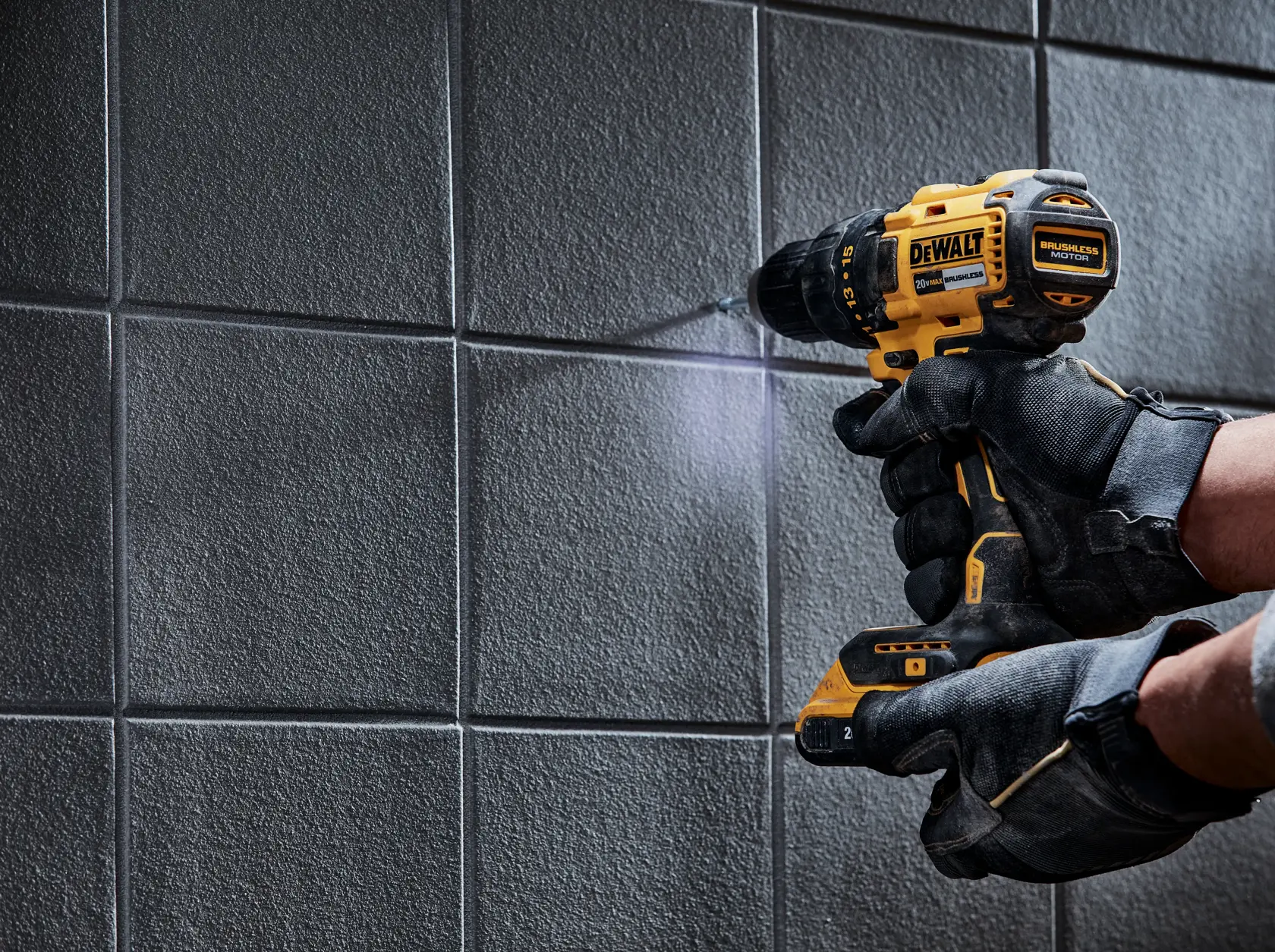 Una persona con guantes negros usa un taladro inalámbrico amarillo DEWALT para perforar una pared de azulejos.