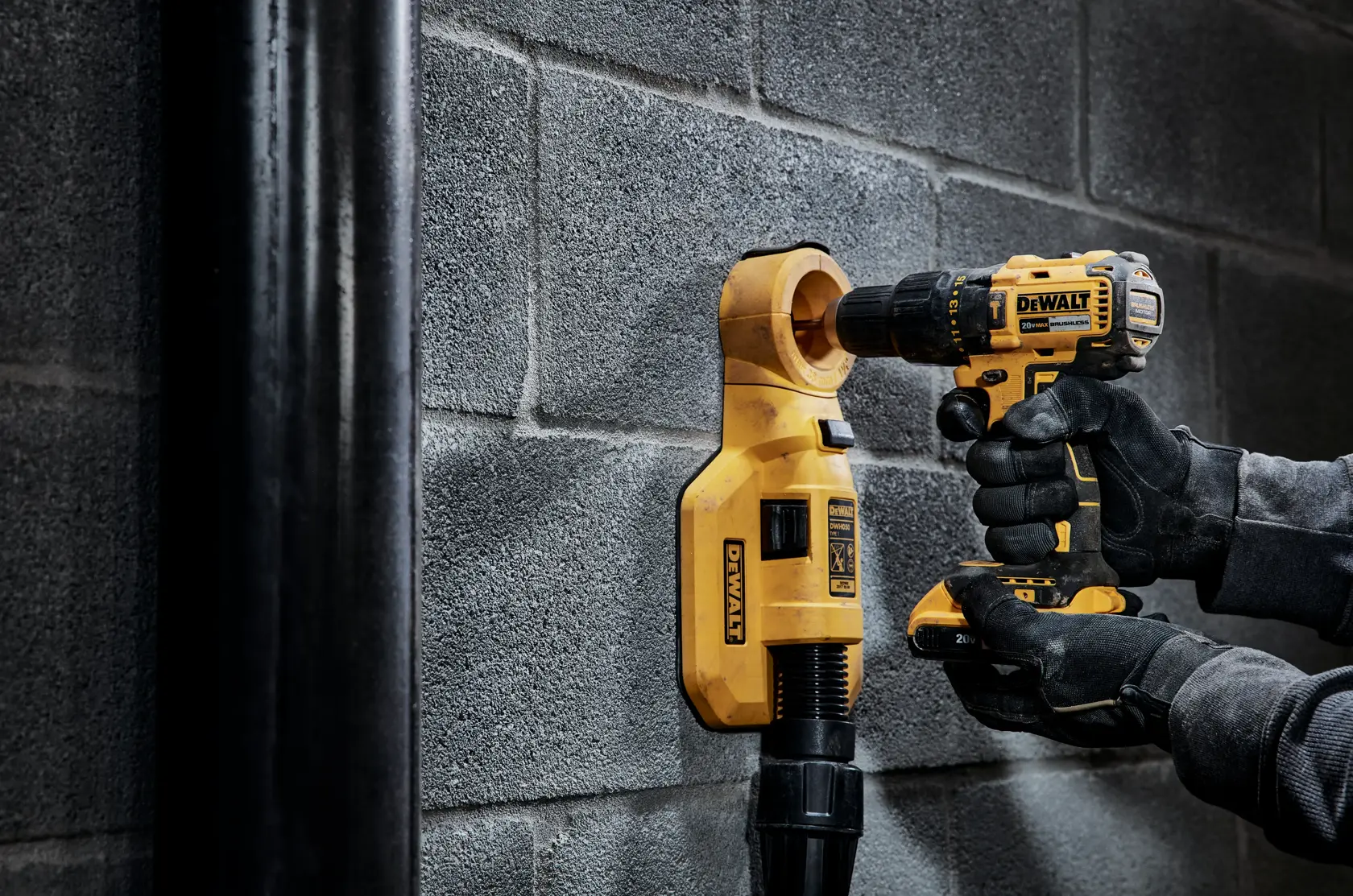 Una persona utiliza un taladro inalámbrico DEWALT en una pared de bloques de hormigón gris con un accesorio de extracción de polvo amarillo DEWALT conectado a una manguera de aspiradora.
