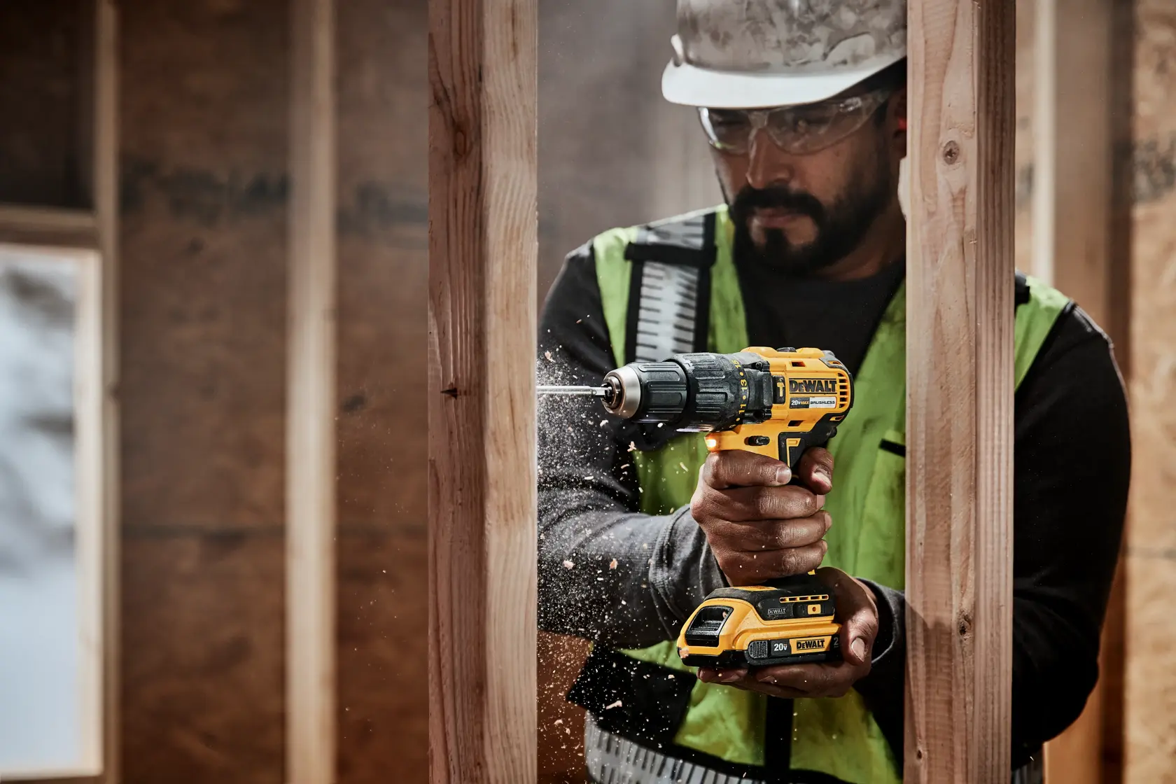 Una persona con chaleco de seguridad y casco está usando un taladro inalámbrico amarillo DEWALT para perforar una viga de madera en un sitio de construcción. La cara de la persona está difuminada.