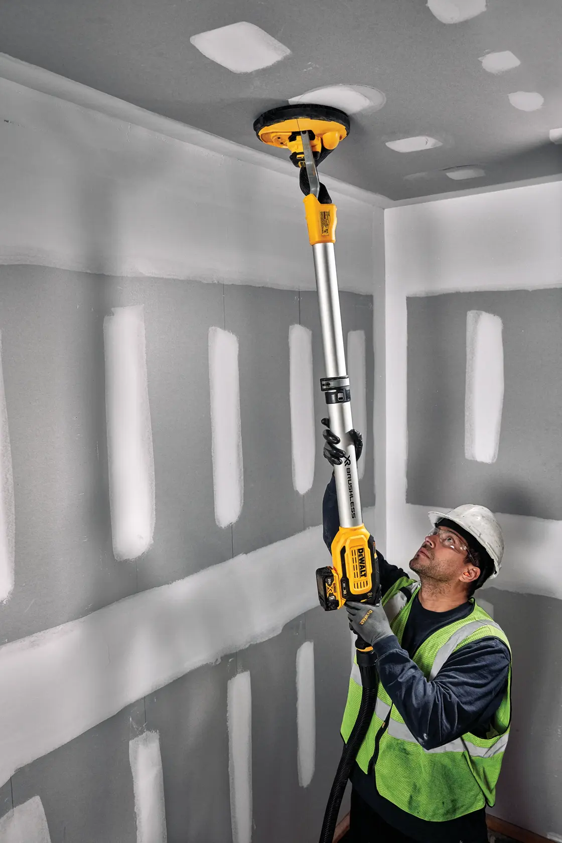 Una persona con chaleco de seguridad y casco usa una lijadora para drywall DEWALT DCE800P2 para lijar el techo en una habitación con drywall sin terminar.