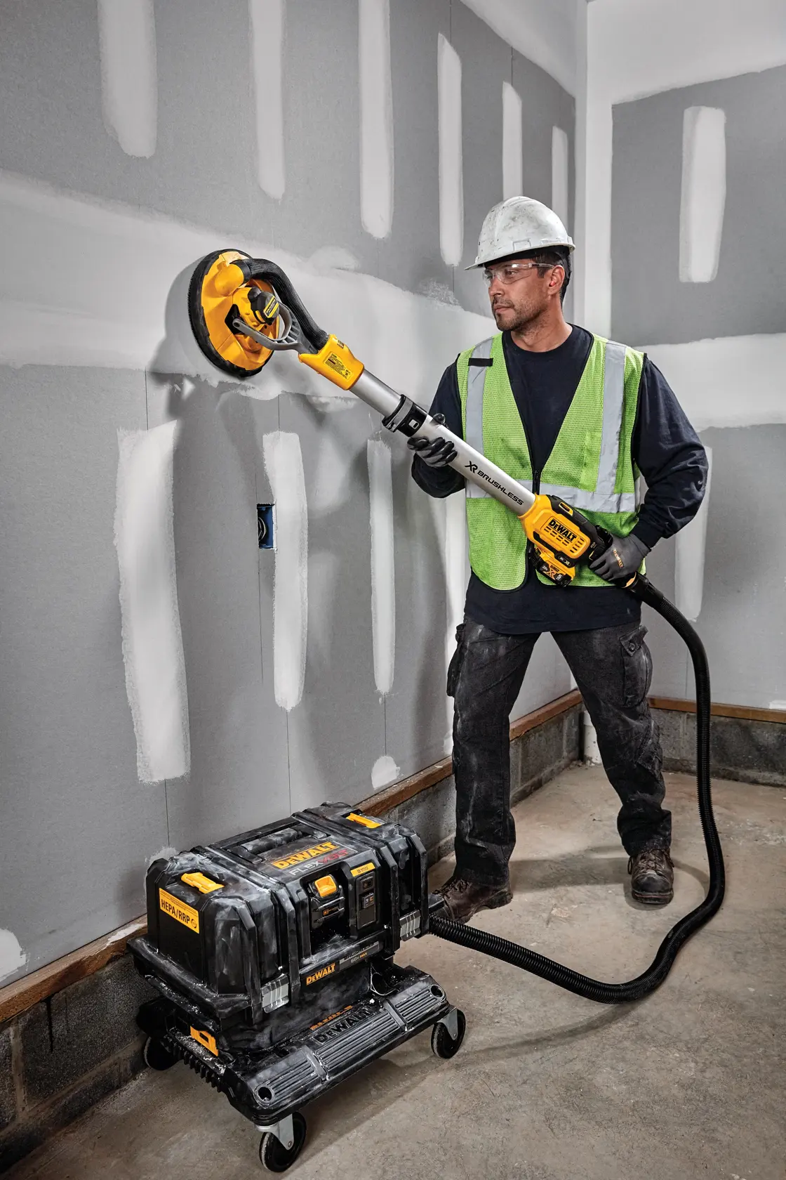 Un trabajador de la construcción con casco de seguridad y chaleco reflectante utiliza una lijadora de drywall DEWALT conectada a un sistema portátil de extracción de polvo. El trabajador está lijando paneles de yeso en una habitación con paredes sin terminar.