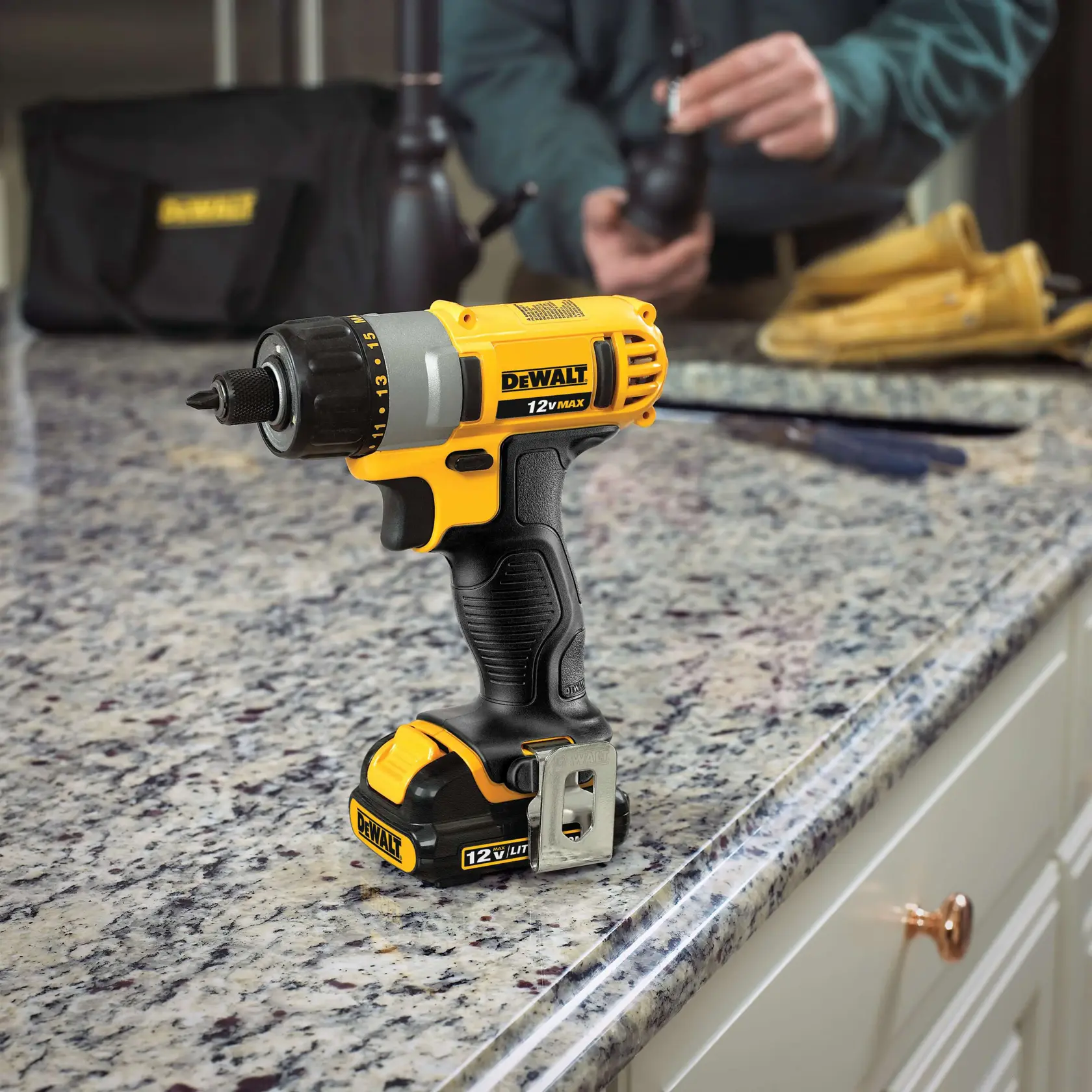 Un taladro inalámbrico DEWALT 12V MAX con batería, de pie sobre una encimera de granito en una cocina. En el fondo, una persona trabaja con herramientas y guantes.