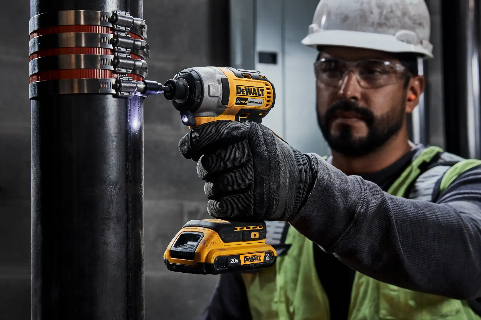 Una persona con guantes de seguridad y casco usa un taladro inalámbrico amarillo DEWALT para apretar abrazaderas metálicas alrededor de una tubería negra grande. El fondo muestra un entorno industrial.
