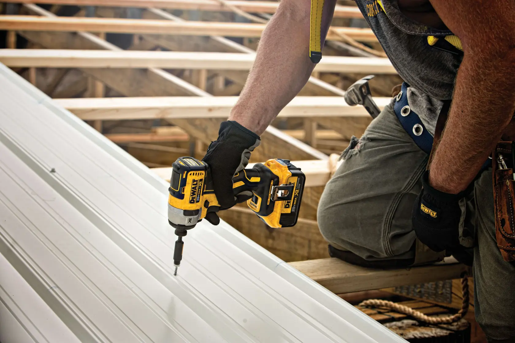 Una persona con guantes de trabajo utiliza un destornillador de impacto inalámbrico DEWALT amarillo y negro para atornillar tornillos en una hoja de metal en un sitio de construcción. Vigas de madera y herramientas son visibles al fondo.
