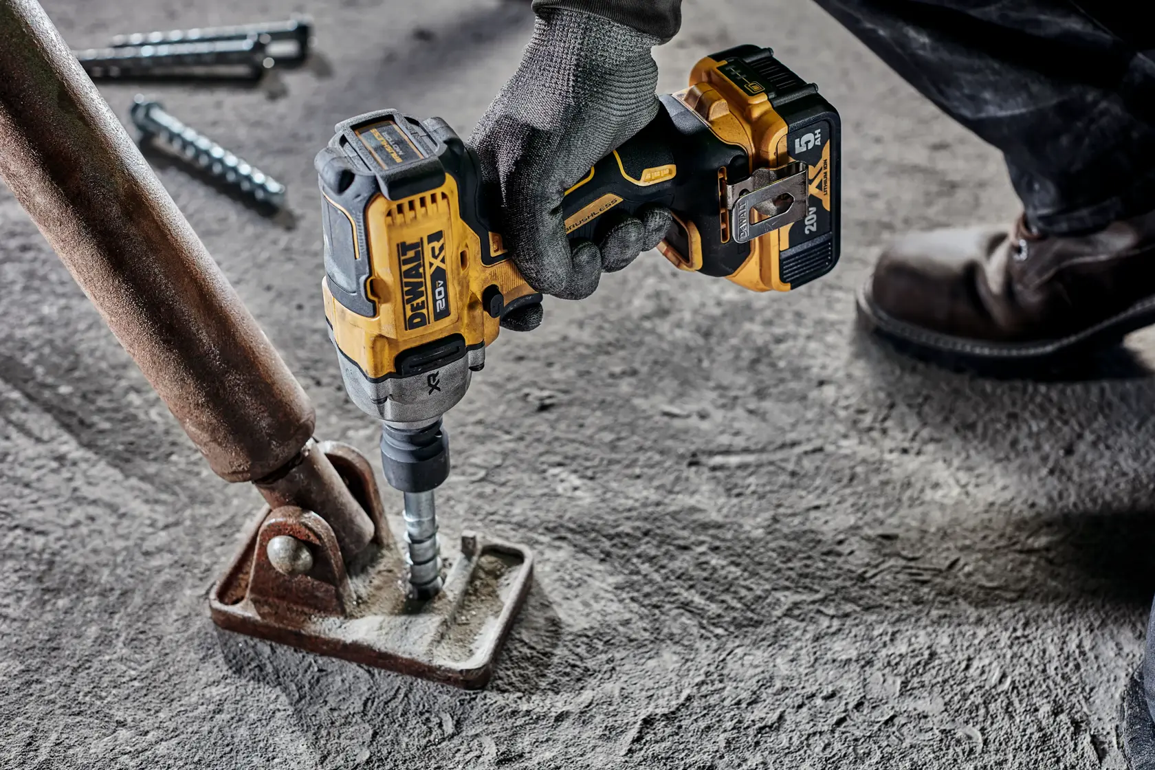 Una persona que lleva guantes de trabajo utiliza una llave de impacto inalámbrica DEWALT 20V (modelo DCF891B) para fijar un perno en una base de metal sobre un suelo de concreto.
