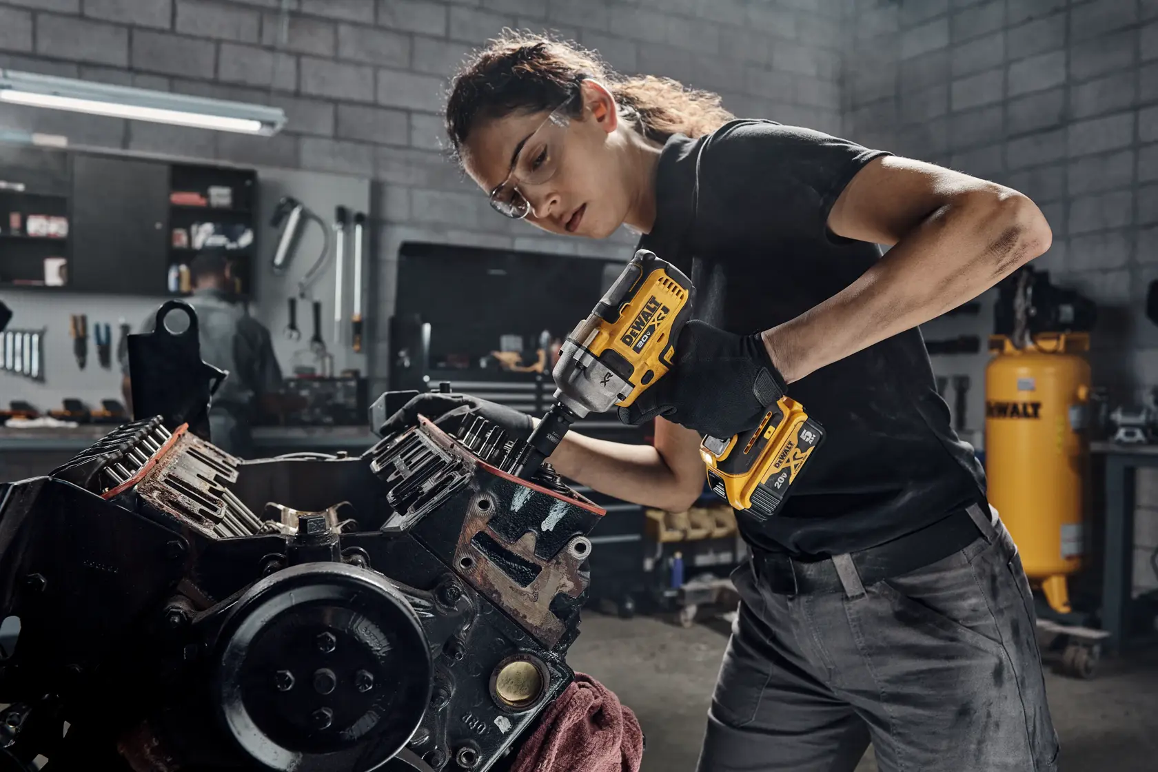 Persona usando una llave de impacto inalámbrica DEWALT DCF891B para trabajar en un motor en un taller. Su rostro está difuminado por privacidad. El fondo del taller incluye herramientas y un compresor de aire DEWALT.
