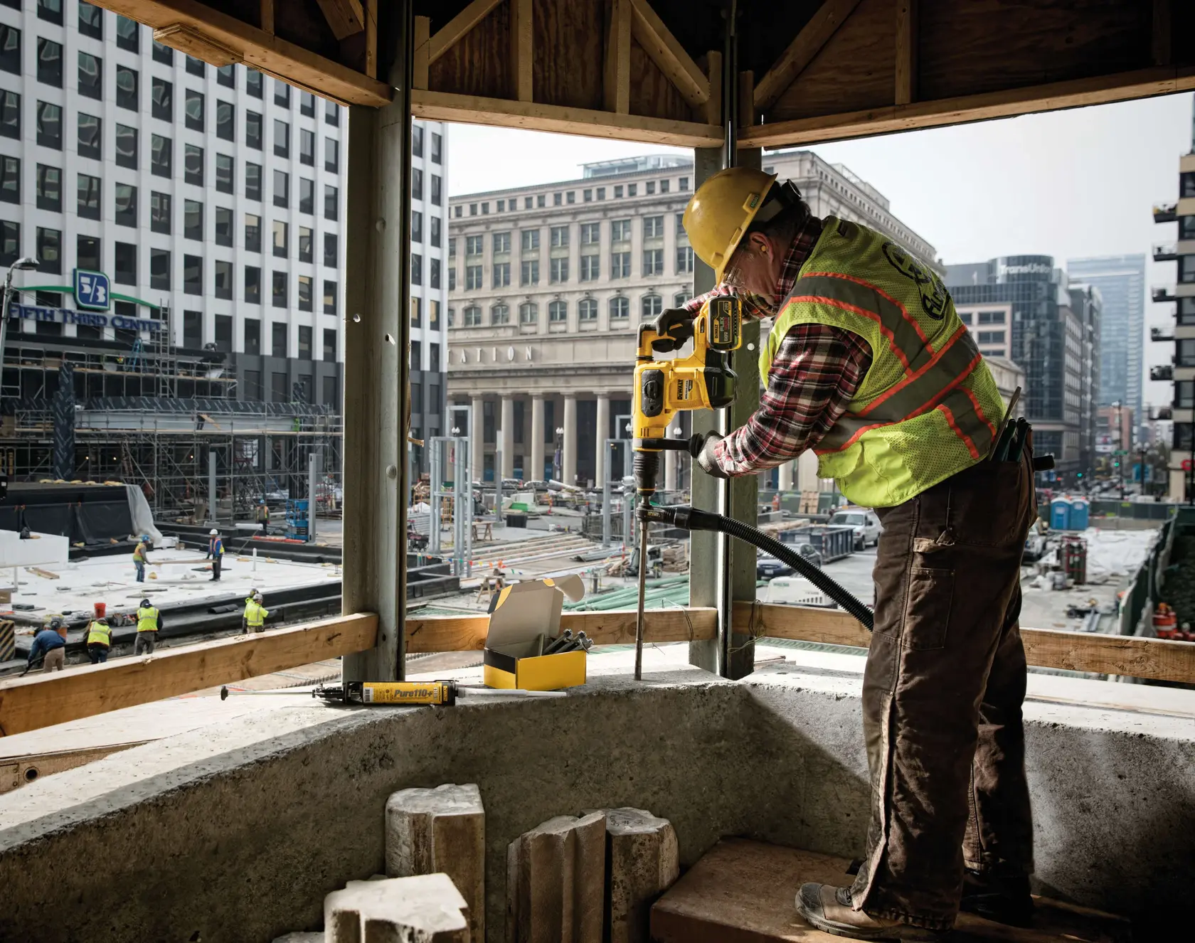 Un trabajador de la construcción con chaleco de seguridad y casco está utilizando una herramienta eléctrica DEWALT para perforar una estructura de concreto en una obra ubicada en una zona urbana. Se pueden ver otros trabajadores y andamios en el fondo.