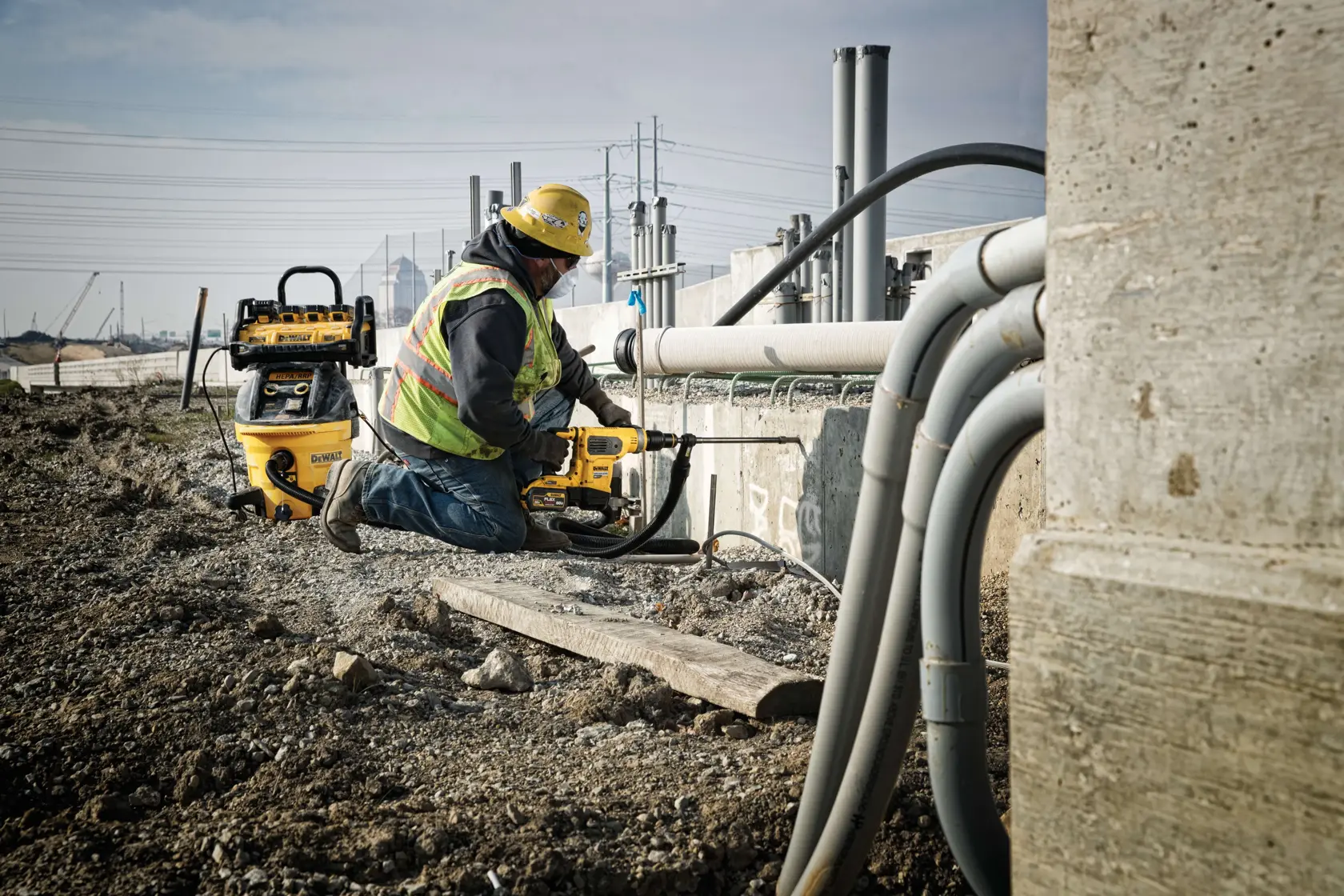 Un trabajador de la construcción con chaleco de seguridad y casco utiliza una herramienta eléctrica amarilla DEWALT en un sitio industrial al aire libre rodeado de tuberías y estructuras de concreto.