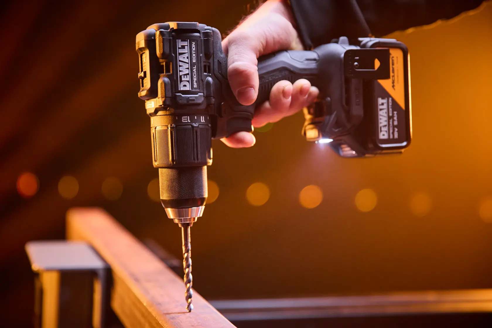 Mano sosteniendo un taladro inalámbrico DEWALT, perforando una superficie de metal con luz de fondo naranja.