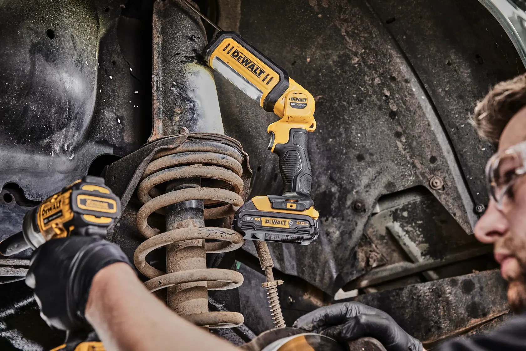Una luz de trabajo LED DEWALT DCL050 se usa para iluminar un sistema de suspensión automotriz mientras una persona con gafas de seguridad y guantes trabaja con otra herramienta DEWALT.