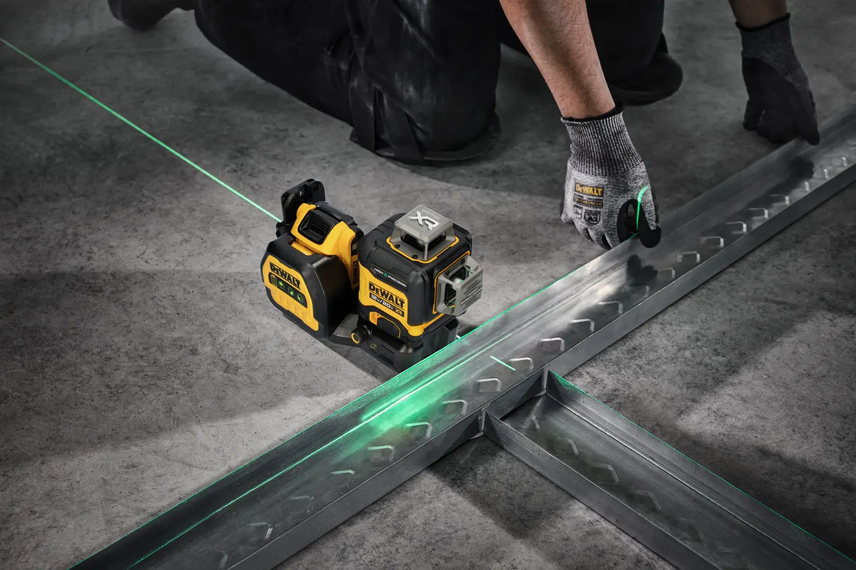 Un dispositivo nivel láser DeWalt proyecta una línea láser verde sobre el suelo de concreto, ayudando a un trabajador con guantes a alinear piezas de estructura metálica.