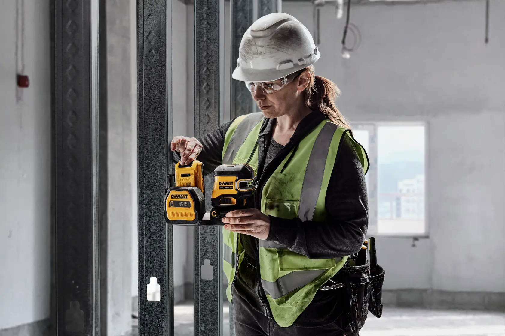 Una persona con casco de seguridad y chaleco de alta visibilidad sostiene un nivel láser DEWALT DCLE34220G de línea cruzada verde en un entorno de construcción.