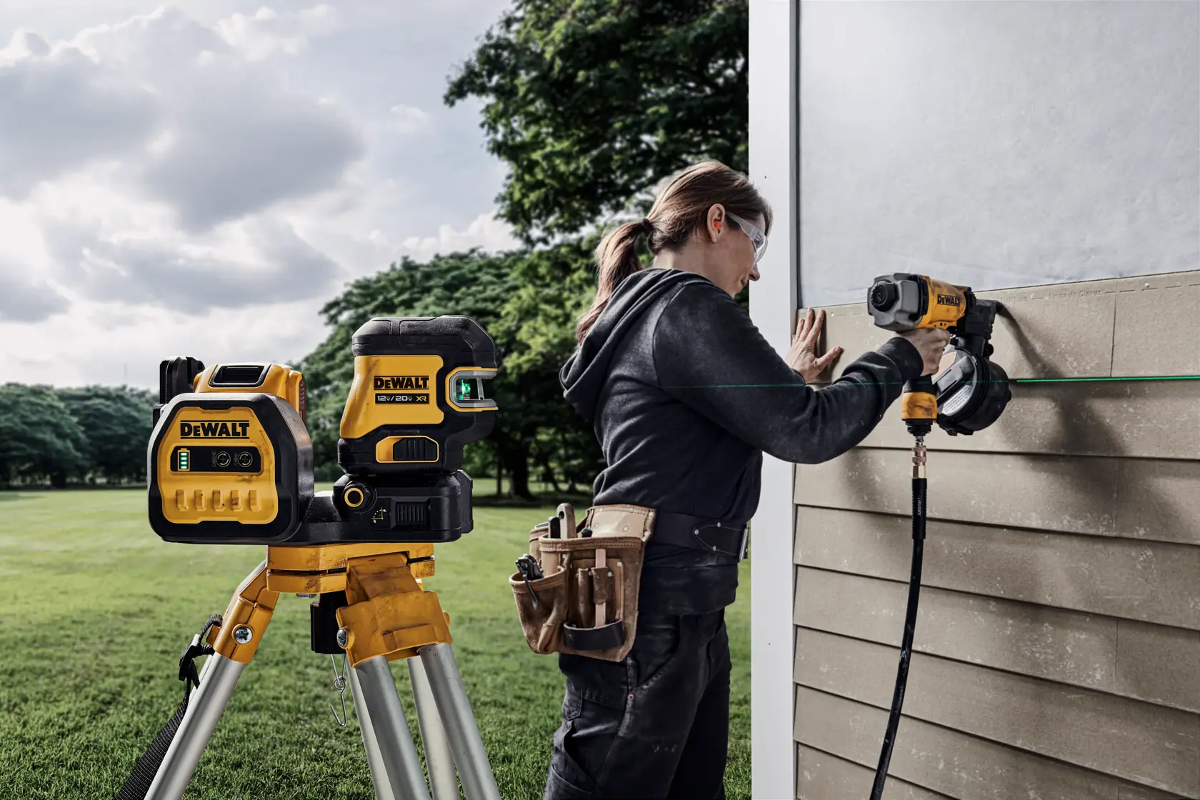 Una persona usando herramientas DEWALT, incluido un nivel láser DEWALT en un trípode, para instalar paneles de revestimiento en una pared exterior. La persona lleva un cinturón de herramientas y trabaja al aire libre.