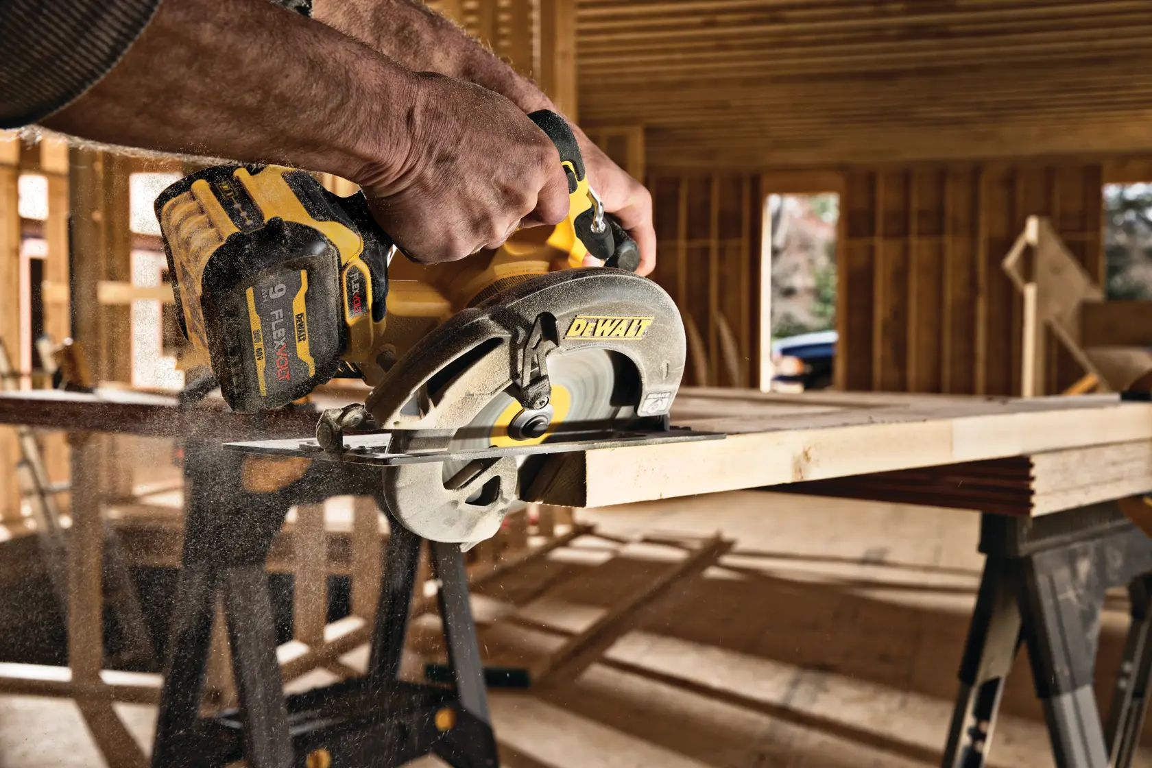 Vista de cerca de una persona usando una sierra circular inalámbrica DEWALT para cortar una tabla de madera dentro de un sitio de construcción. Se puede ver polvo de madera y el fondo muestra estructuras de madera.