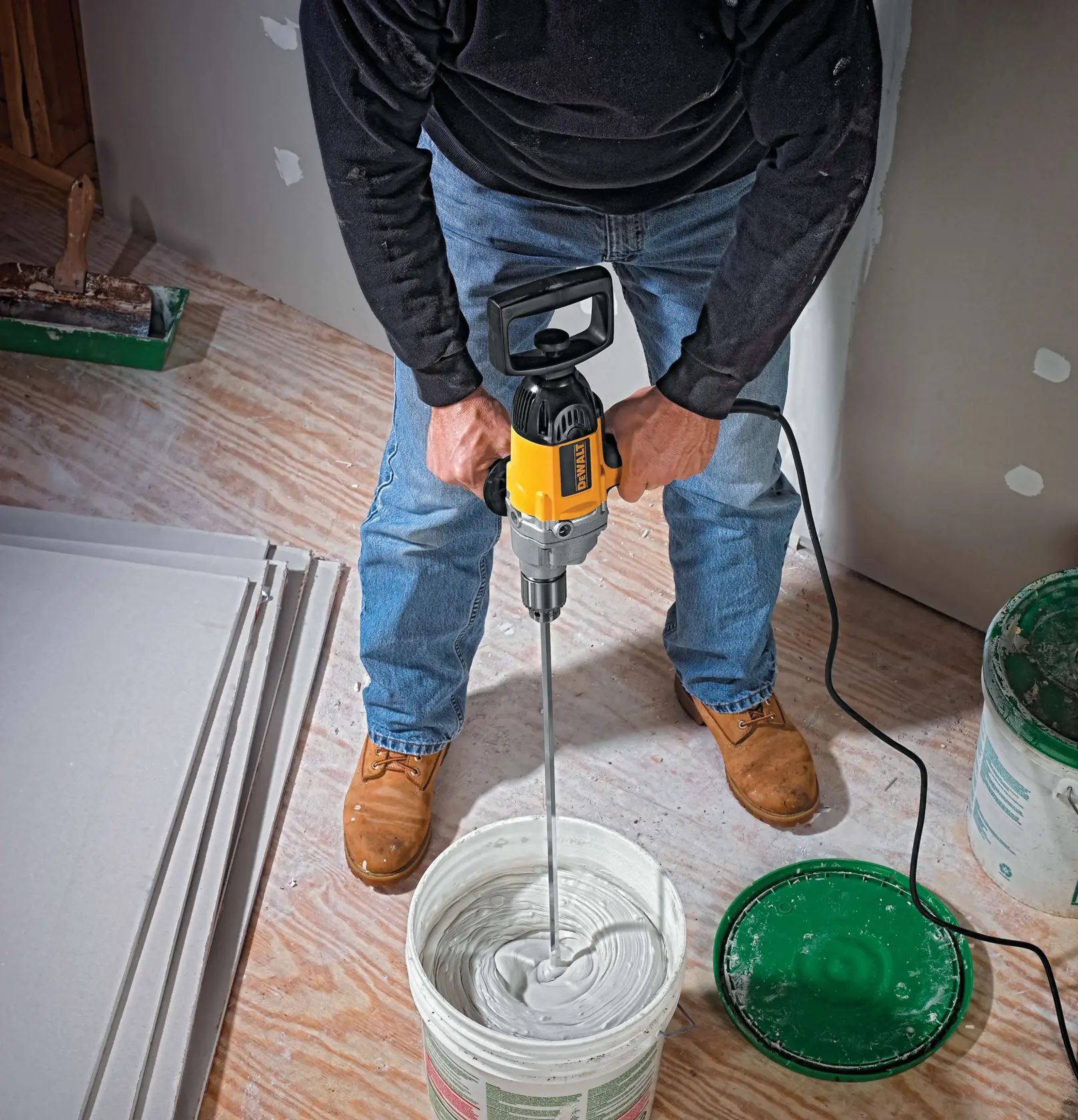 Una persona utiliza un taladro eléctrico DEWALT DW130V para mezclar yeso o pasta de juntas en un balde grande, con paneles de yeso y herramientas visibles en el suelo.