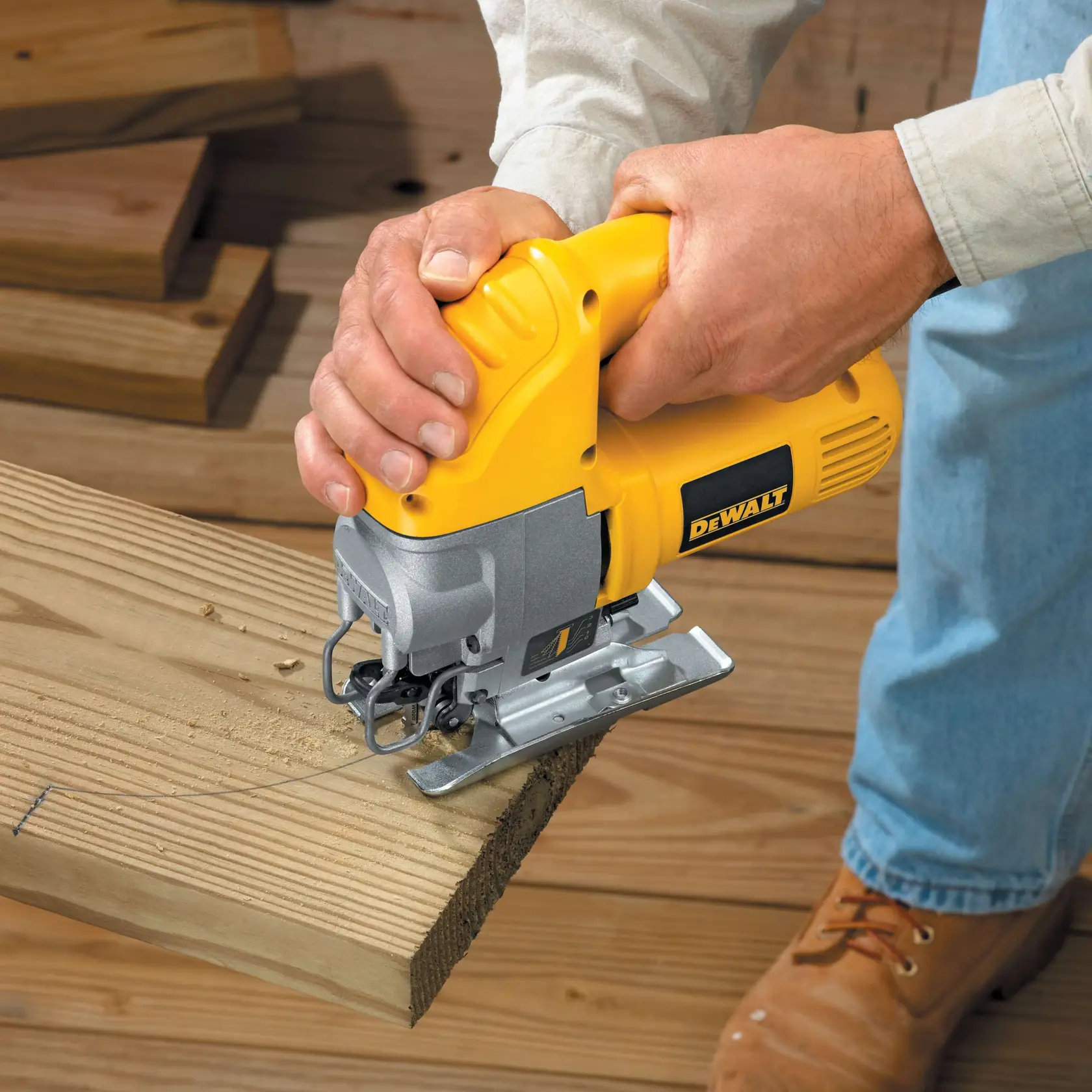 Una persona está usando una sierra de calar amarilla DEWALT para cortar un trozo de madera. La herramienta se sostiene firmemente y está realizando un corte curvado en la tabla. La persona lleva una camisa de manga larga, pantalones vaqueros y botas de trabajo.