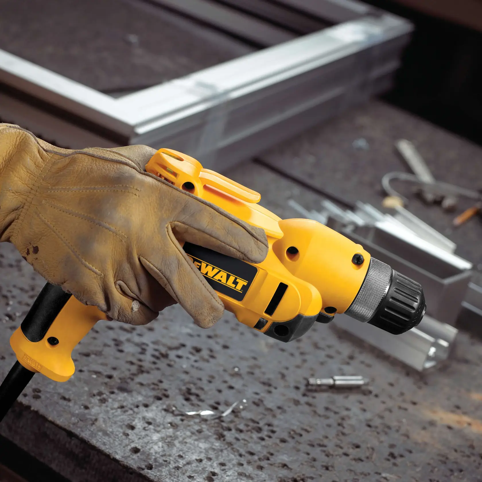 Una mano con guante sostiene un taladro con cable amarillo DEWALT, colocado sobre una mesa de trabajo con piezas de metal y herramientas en el fondo.