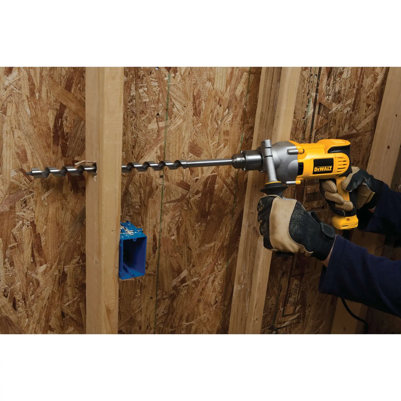Una persona con guantes de trabajo usa un taladro eléctrico amarillo DEWALT para perforar un agujero grande en una viga de madera dentro de una estructura de construcción. El taladro tiene una broca larga tipo espiral y cerca hay una caja de toma eléctrica azul montada.