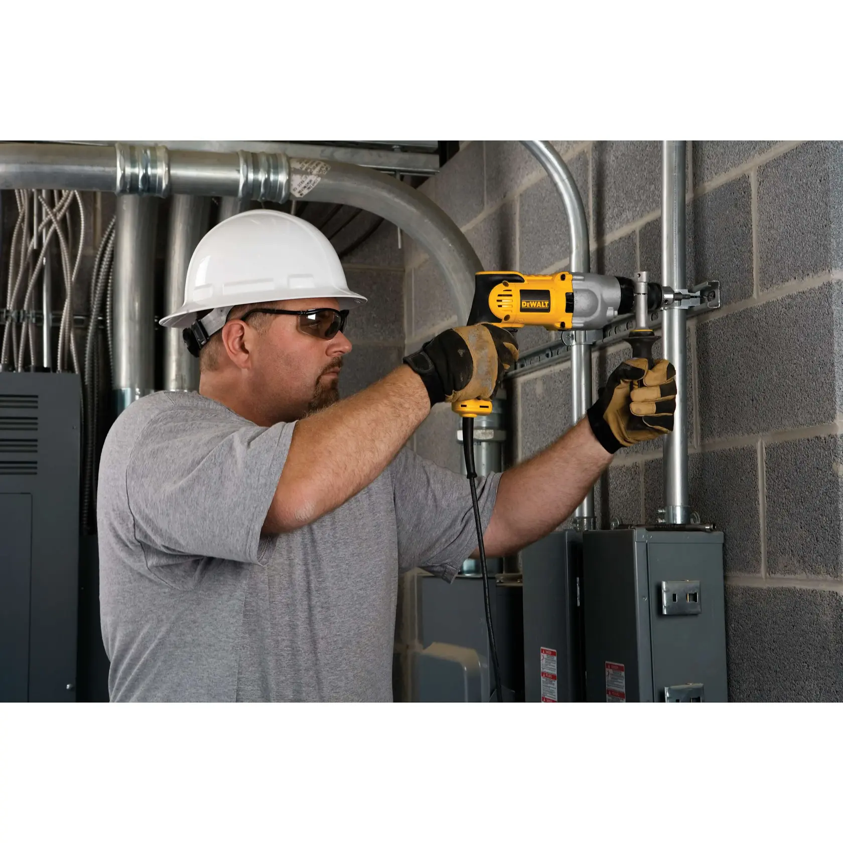Una persona con casco de seguridad y guantes utiliza un taladro con cable DEWALT DWD520K para instalar o asegurar tuberías metálicas en la pared de una sala industrial o de servicios.