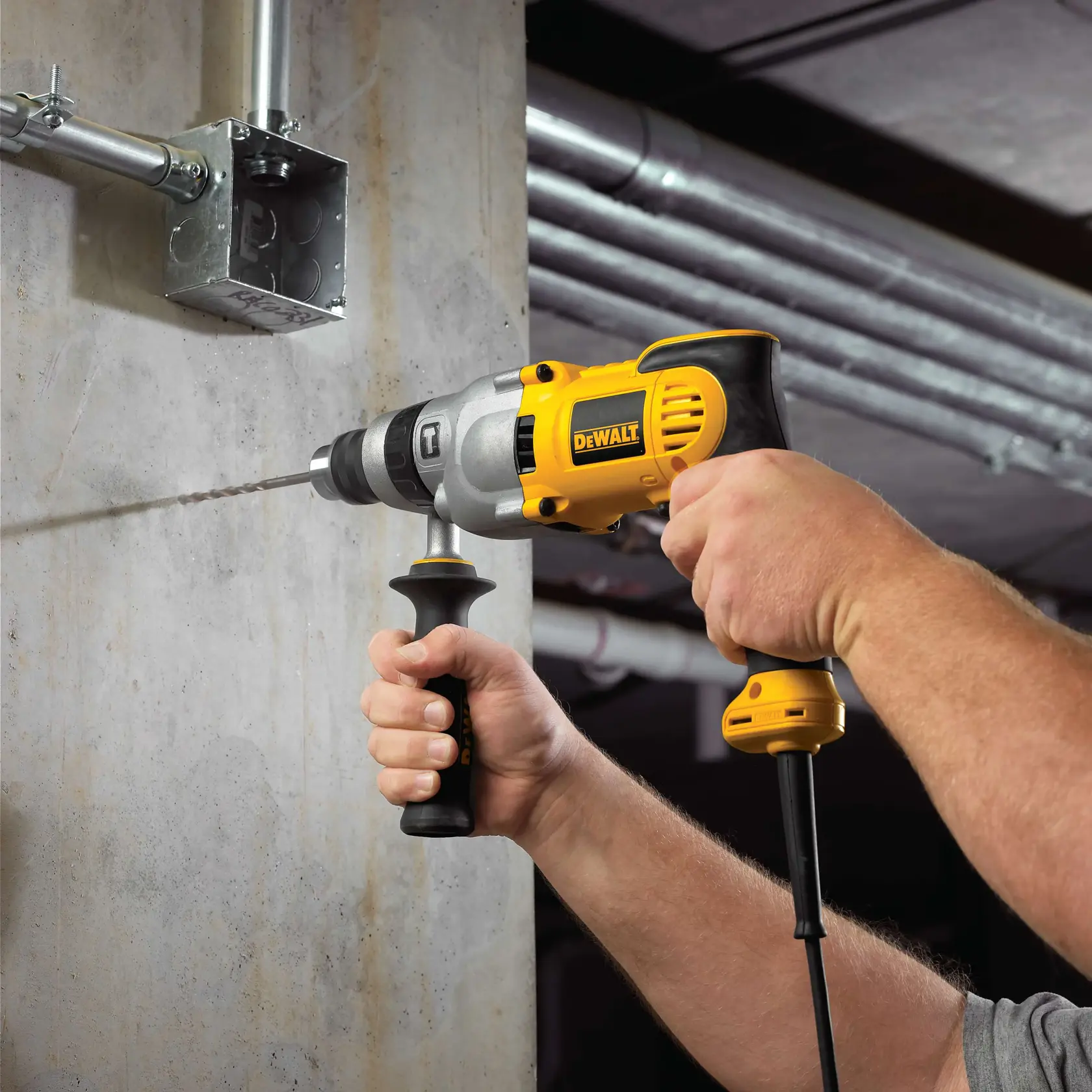 Una persona utiliza un taladro eléctrico DEWALT DWD520 para perforar un agujero en una pared de hormigón cerca de tubos metálicos y una caja eléctrica.
