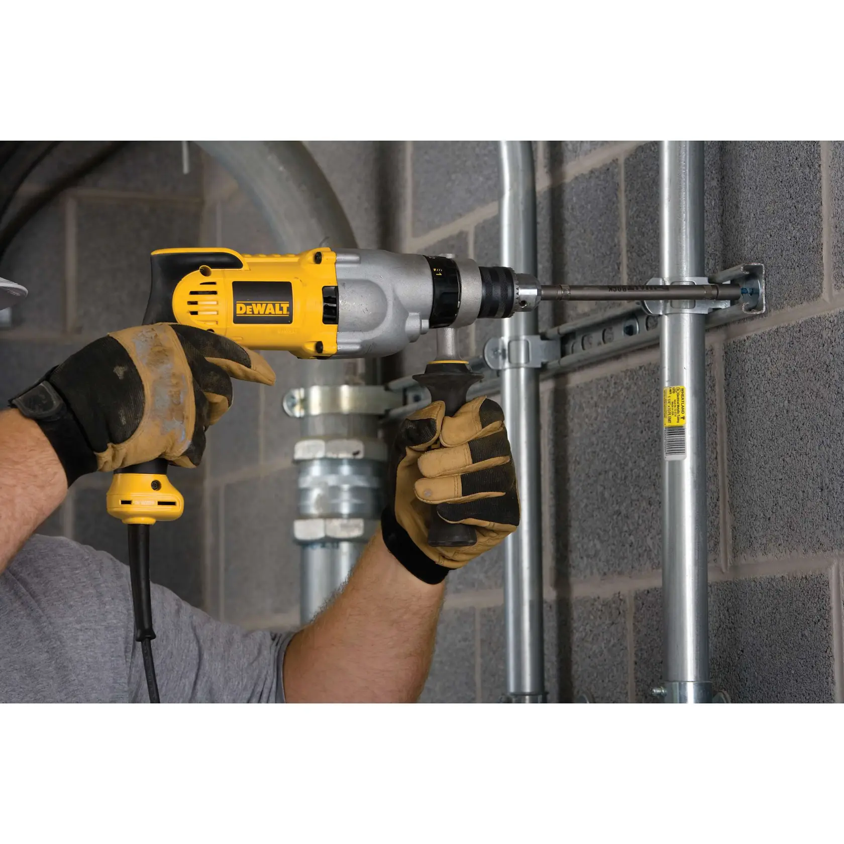 Una persona con guantes de trabajo utiliza un taladro con cable amarillo y gris DEWALT DWD520 para perforar una pieza metálica montada en una pared de hormigón.