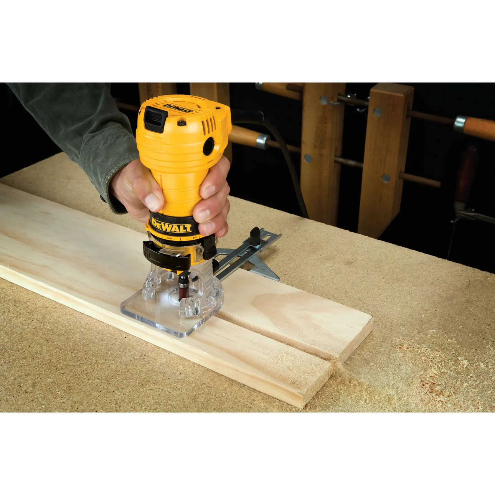 Una persona usando una fresadora para madera DEWALT DWE6000 para tallar o dar forma a una pieza de madera clara sobre una mesa de trabajo. La fresadora es de color amarillo y negro, y está posicionada sobre la madera mientras el aserrín se acumula alrededor.