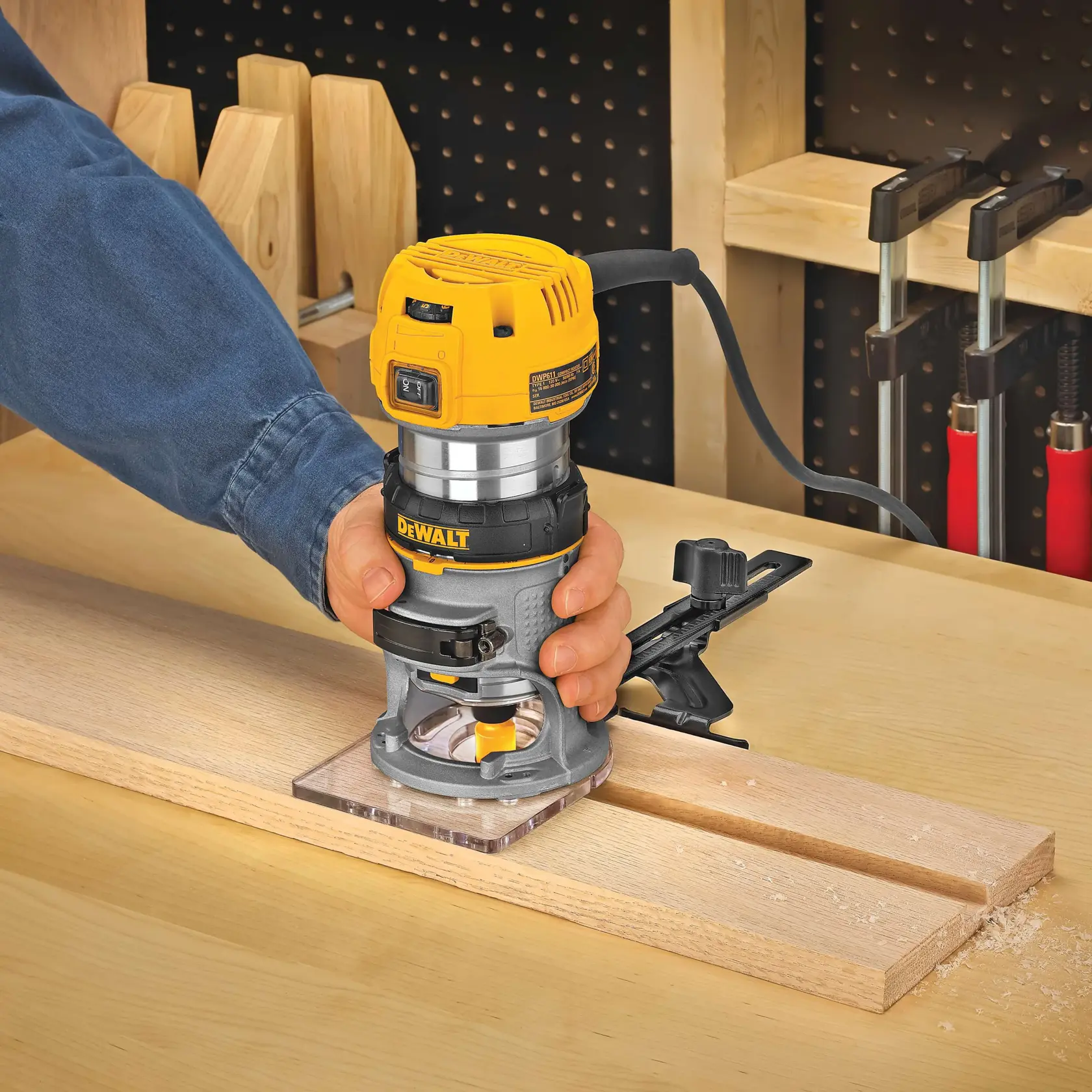 Una persona utiliza una fresadora de madera DEWALT DWP611 para tallar ranuras en una tabla de madera sobre una mesa de trabajo. La fresadora está conectada y fijada para mayor precisión. En el fondo se pueden ver herramientas y abrazaderas de taller.