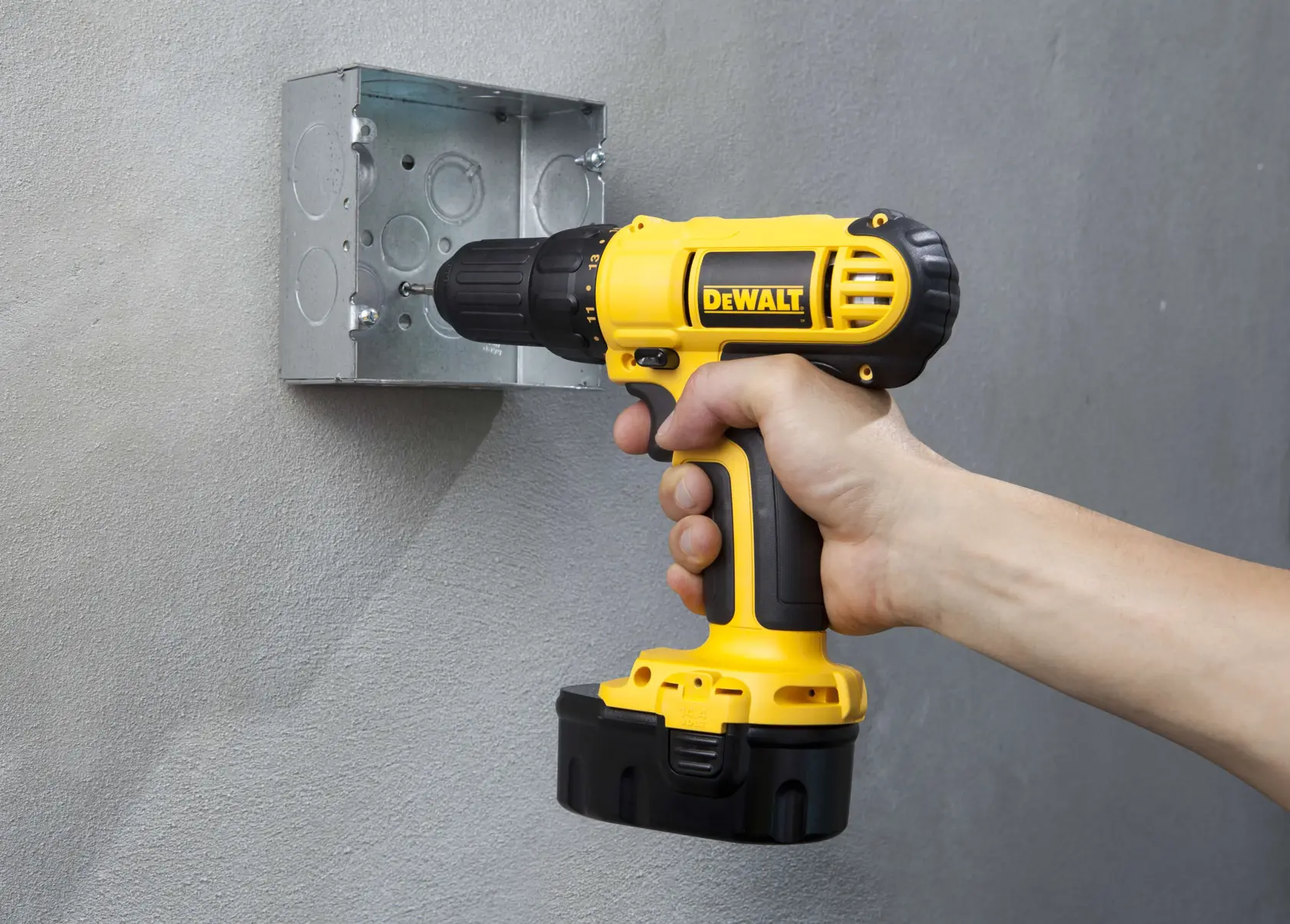 Una mano utiliza un taladro inalámbrico amarillo DEWALT para atornillar o fijar un tornillo en una caja eléctrica de metal montada en una pared gris.