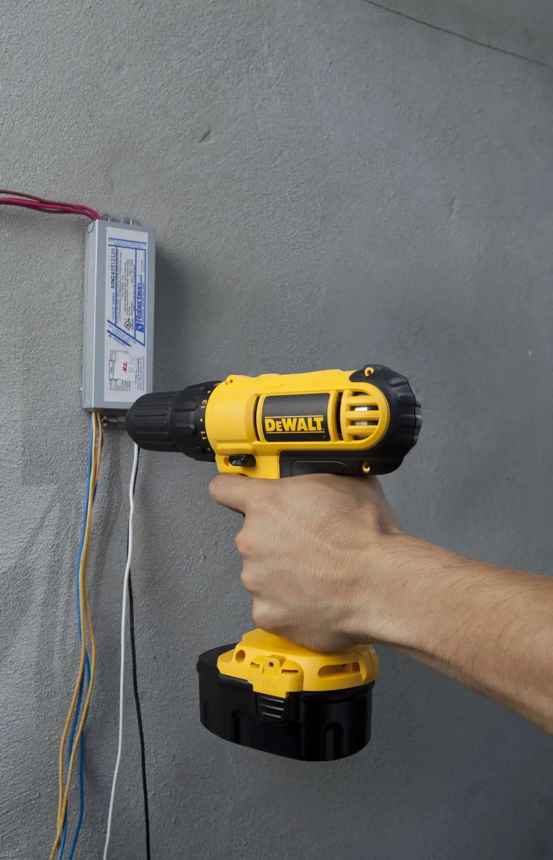 Una mano sostiene un taladro inalámbrico amarillo y negro DEWALT, utilizándolo junto a un componente eléctrico montado en una pared gris con cables conectados.