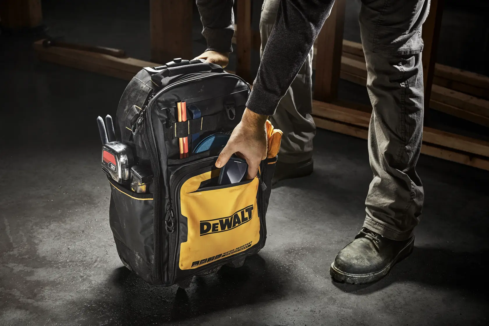 Una persona sostiene una mochila para herramientas DEWALT llena de varias herramientas y accesorios. La mochila es negra con un bolsillo frontal amarillo que muestra el logo de DEWALT. La escena parece estar en un taller o entorno de construcción.