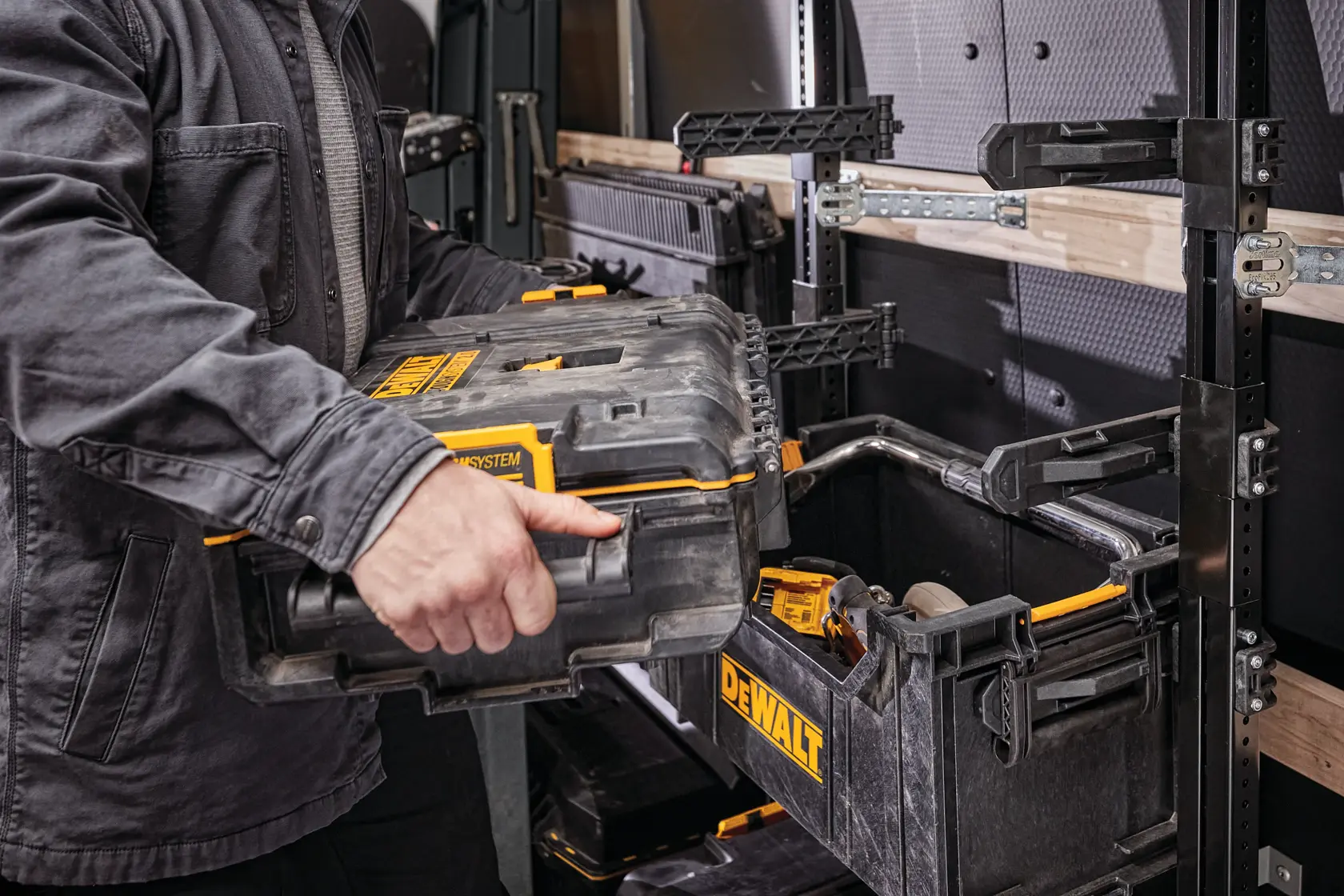 Una persona coloca una caja de herramientas DEWALT ToughSystem en un estante de almacenamiento dentro de un vehículo. Se ven varias cajas y herramientas de almacenamiento DEWALT, y el estante está montado en la pared para organización segura.